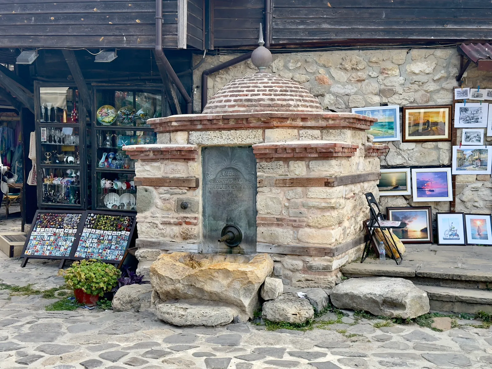 Oude stenen fontein op een geplaveid straatje in Nessebar, omringd door souvenirwinkels en schilderijen