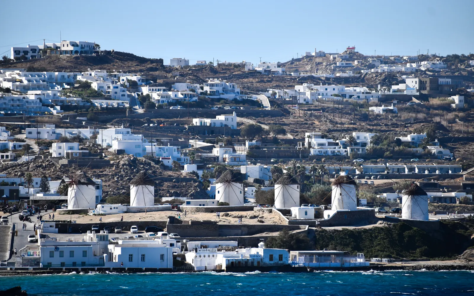 Rij witte windmolens van Mykonos aan zee met daarachter een heuvel vol witte huizen