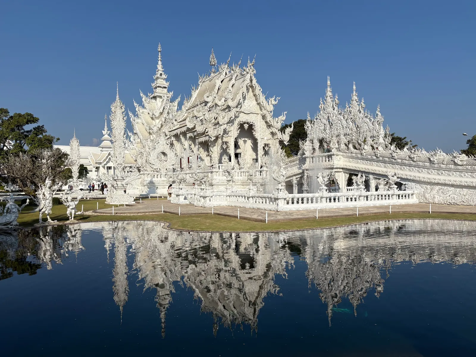 De spierwitte Wat Rong Khun in Chiang Rai weerspiegeld in een vijver onder een strakblauwe lucht
