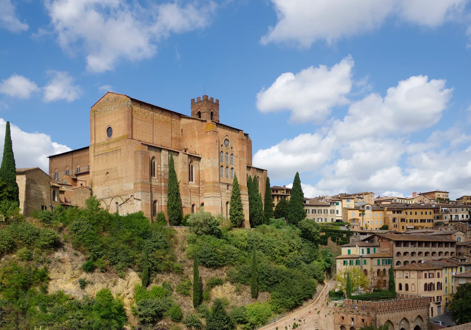 Bakstenen Basiliek van San Domenico op een groene heuvel in Siena onder een blauwe lucht met wolken