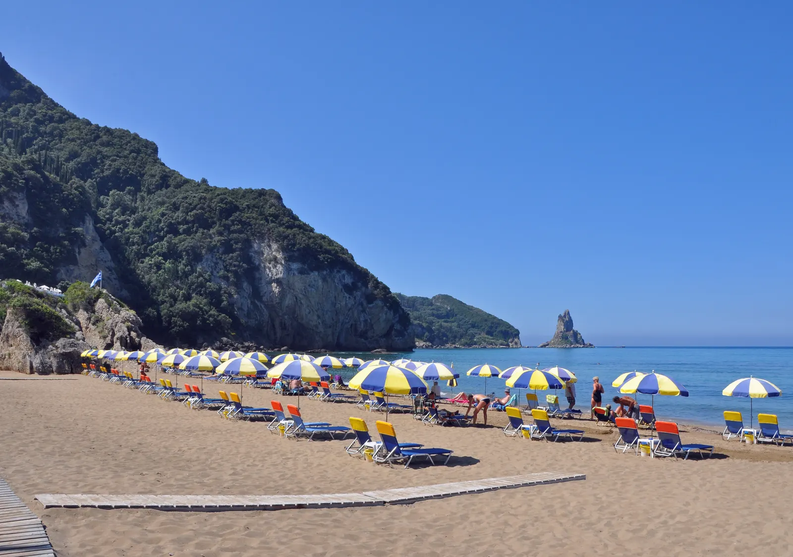 Zandstrand van Agios Gordios met parasols en ligbedden, groene kliffen en de rotspunt Ortholithos in zee