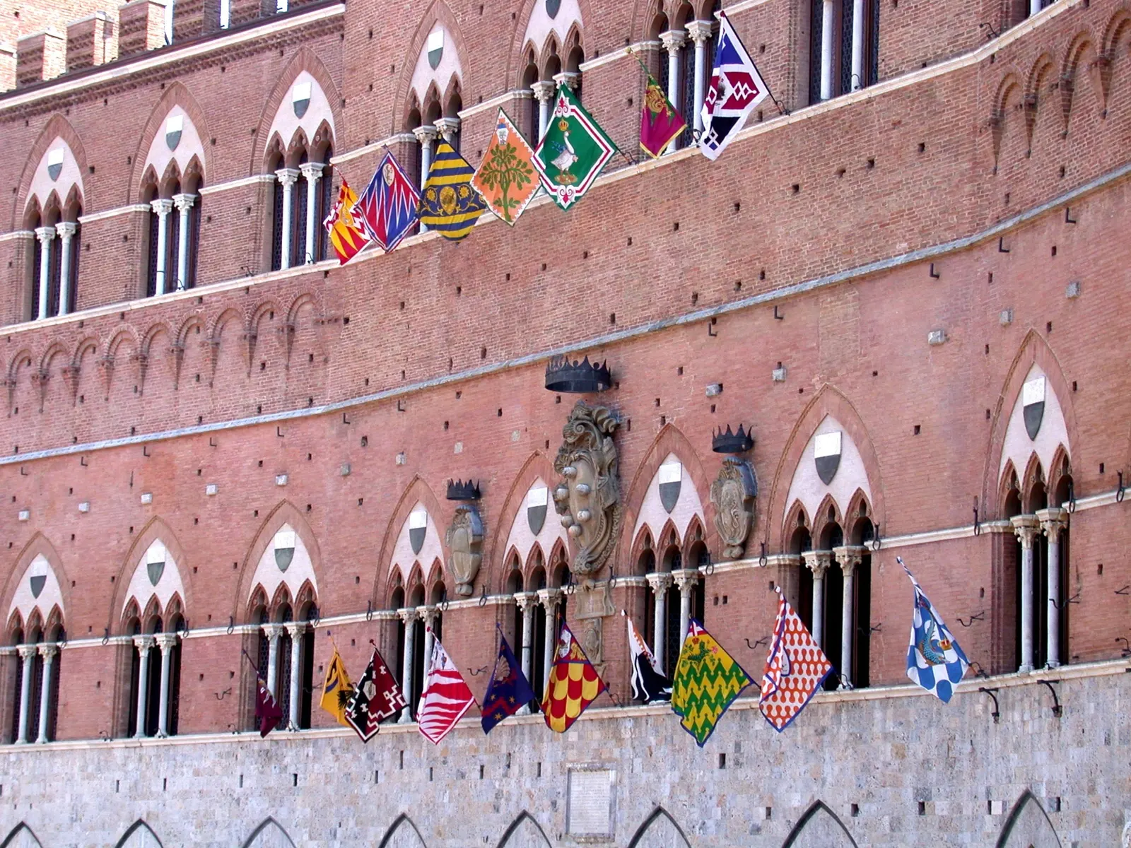 Bakstenen gevel van het Palazzo Pubblico in Siena met gotische ramen en kleurrijke contradevlaggen