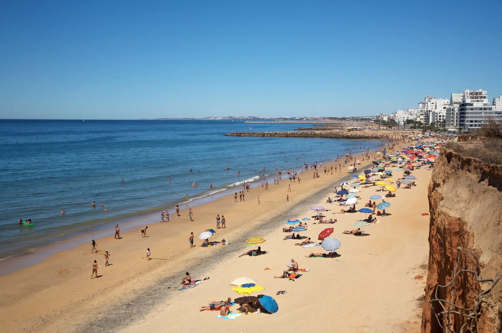 Zonnig zandstrand met parasols en zwemmers, rode kliffen en appartementen aan de kust van Quarteira
