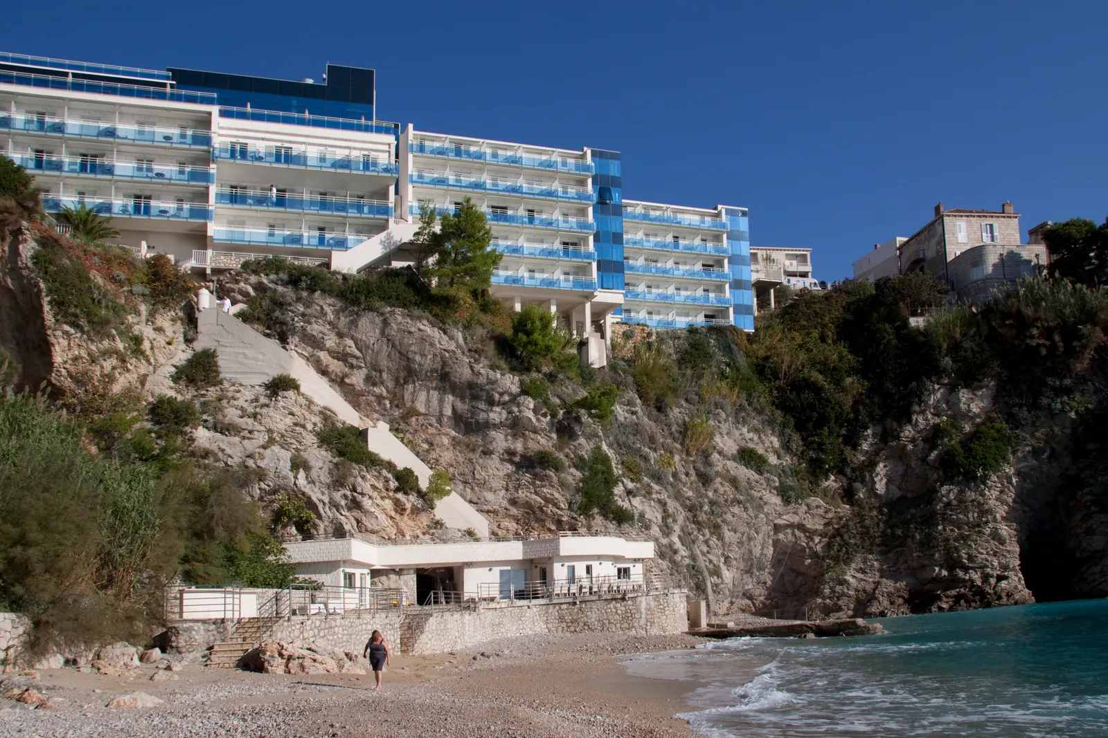 Hotel Bellevue Dubrovnik met blauwe balkons boven op de rotsklif, met een kiezelstrand en zee eronder