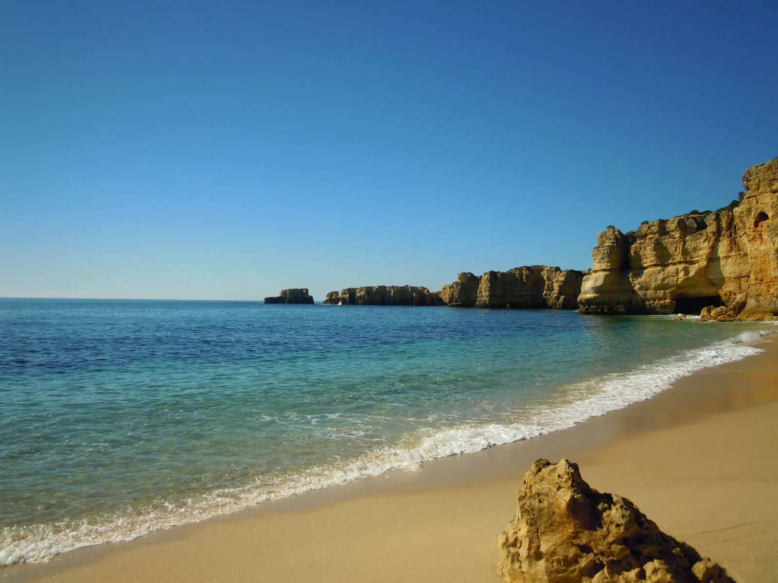 Zandstrand met turquoise zee en goudkleurige kliffen onder een strakblauwe lucht aan de Algarve-kust