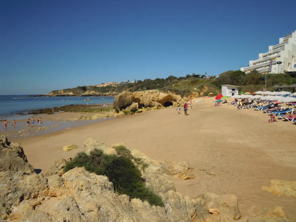 Zandstrand Praia da Oura met rotsen, ligbedden en witte hotels op de klif bij helderblauwe lucht