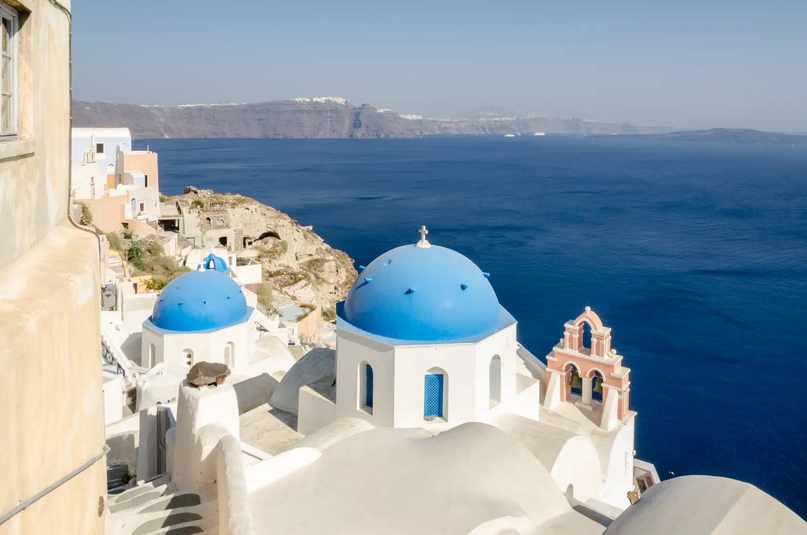 Witte kerkjes met blauwe koepels en een roze klokkentoren boven de diepblauwe caldera van Oia, Santorini