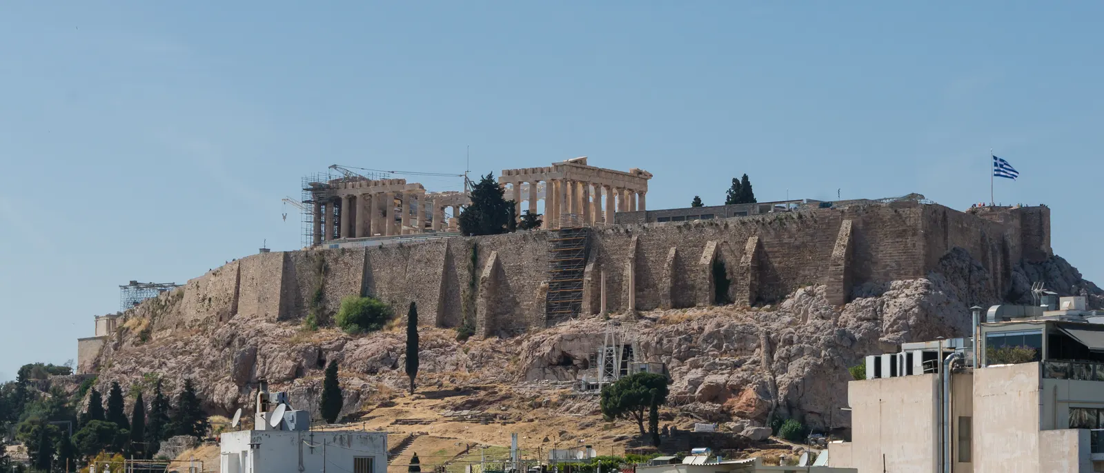 De Akropolis op de rotsheuvel boven Athene met het Parthenon en de Griekse vlag tegen een blauwe lucht