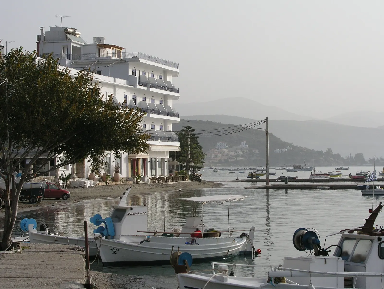 Witte vissersbootjes in het water voor een hotel aan de zeezijde van Tolo, met bergen op de achtergrond