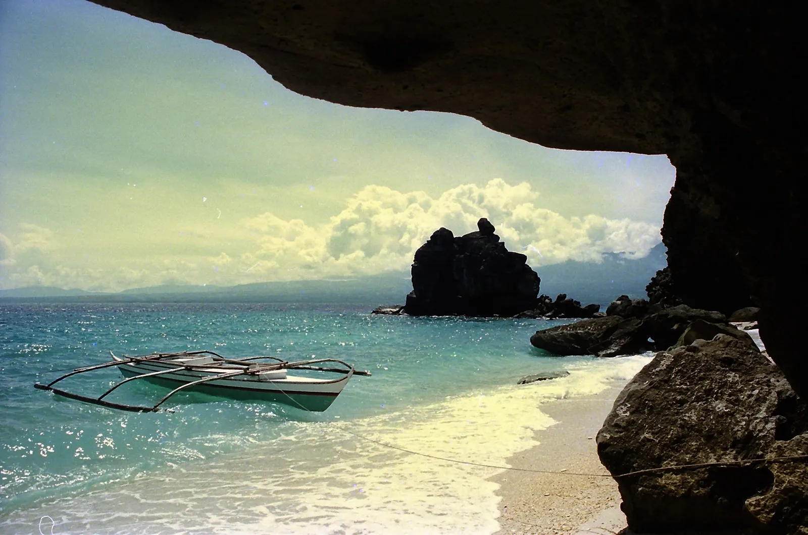 Strand met witte bangka-boot en rotspunt in turquoise water, gezien vanuit een rotsgrot op Apo Island