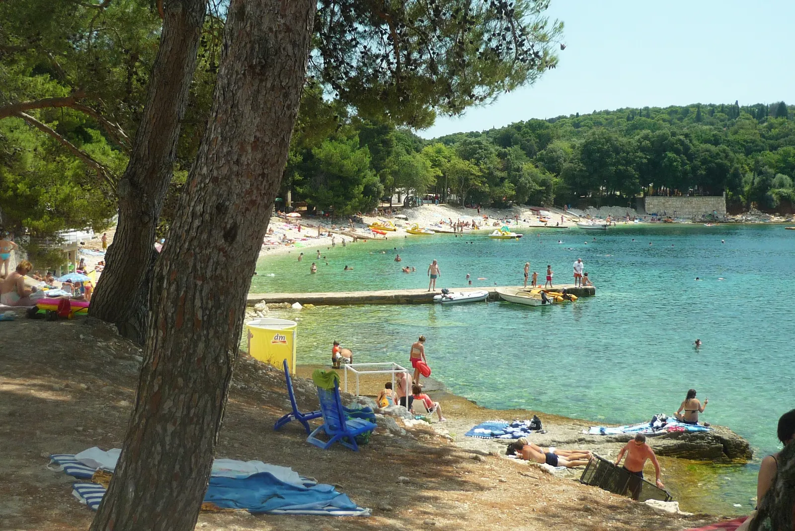 Baai met turquoise water, pijnbomen en een betonnen steiger, met strandgangers op de rotsige kust