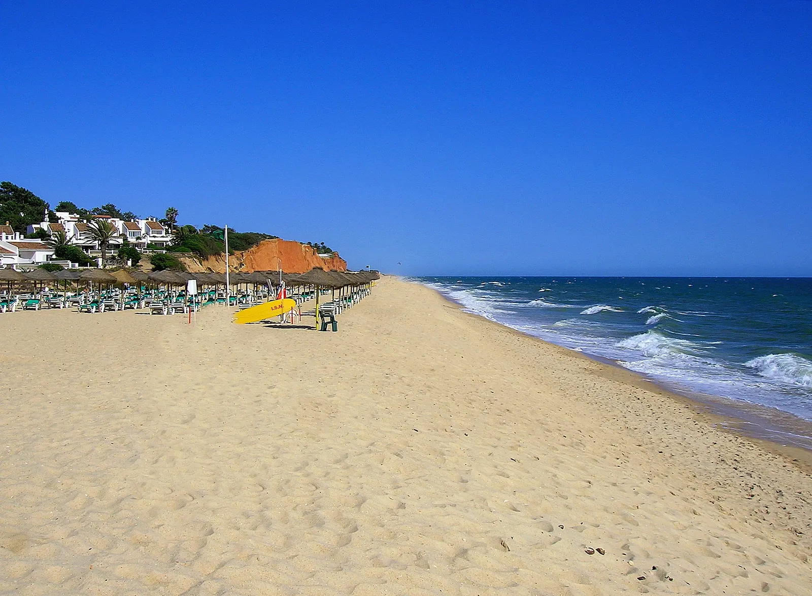 Breed zandstrand van Vale do Lobo met parasols, rode kliffen en golvende blauwe zee onder strakblauwe lucht