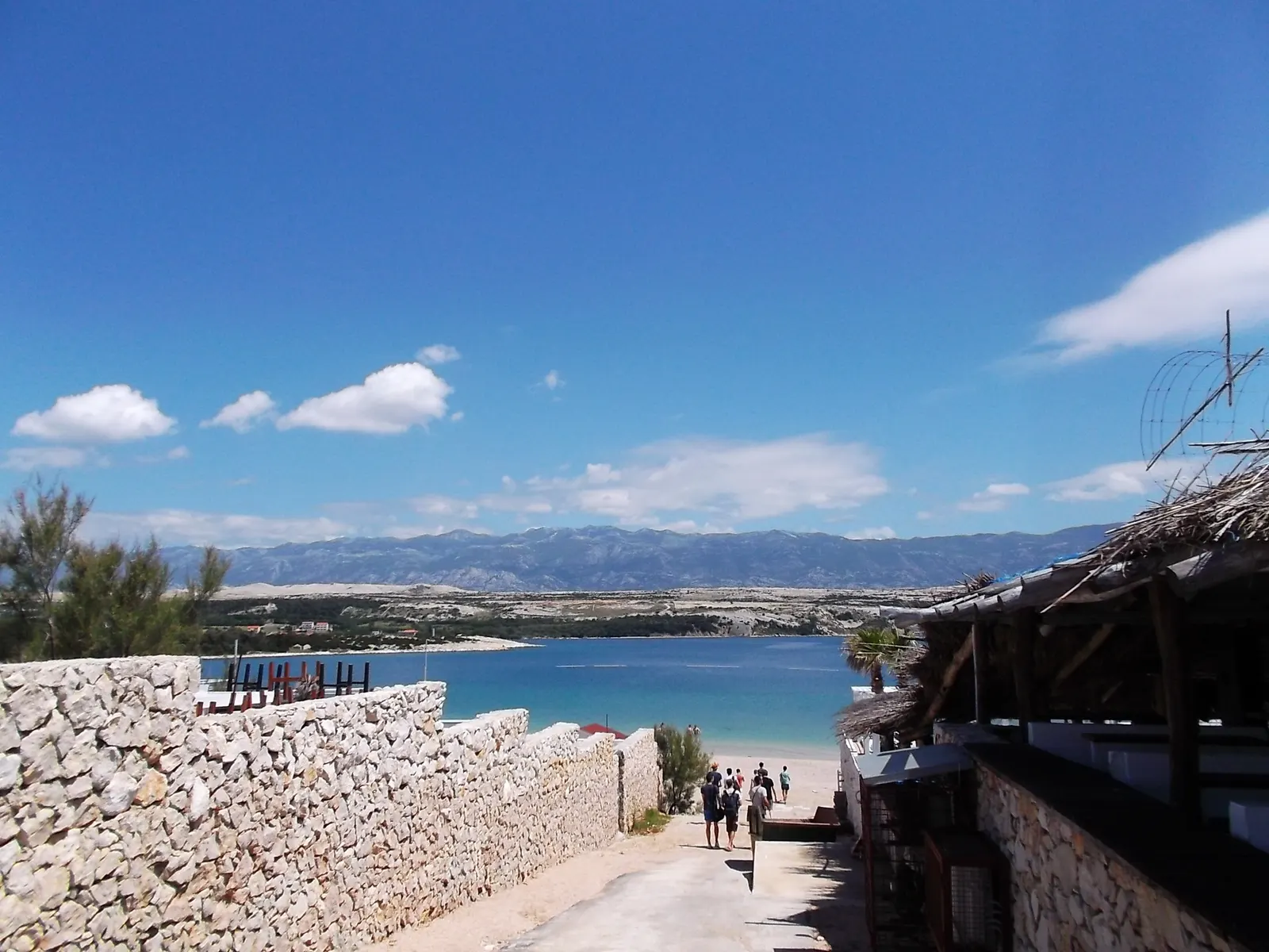 Pad tussen stenen muren naar Zrće strand met turquoise baai en kale bergen van Pag op de achtergrond