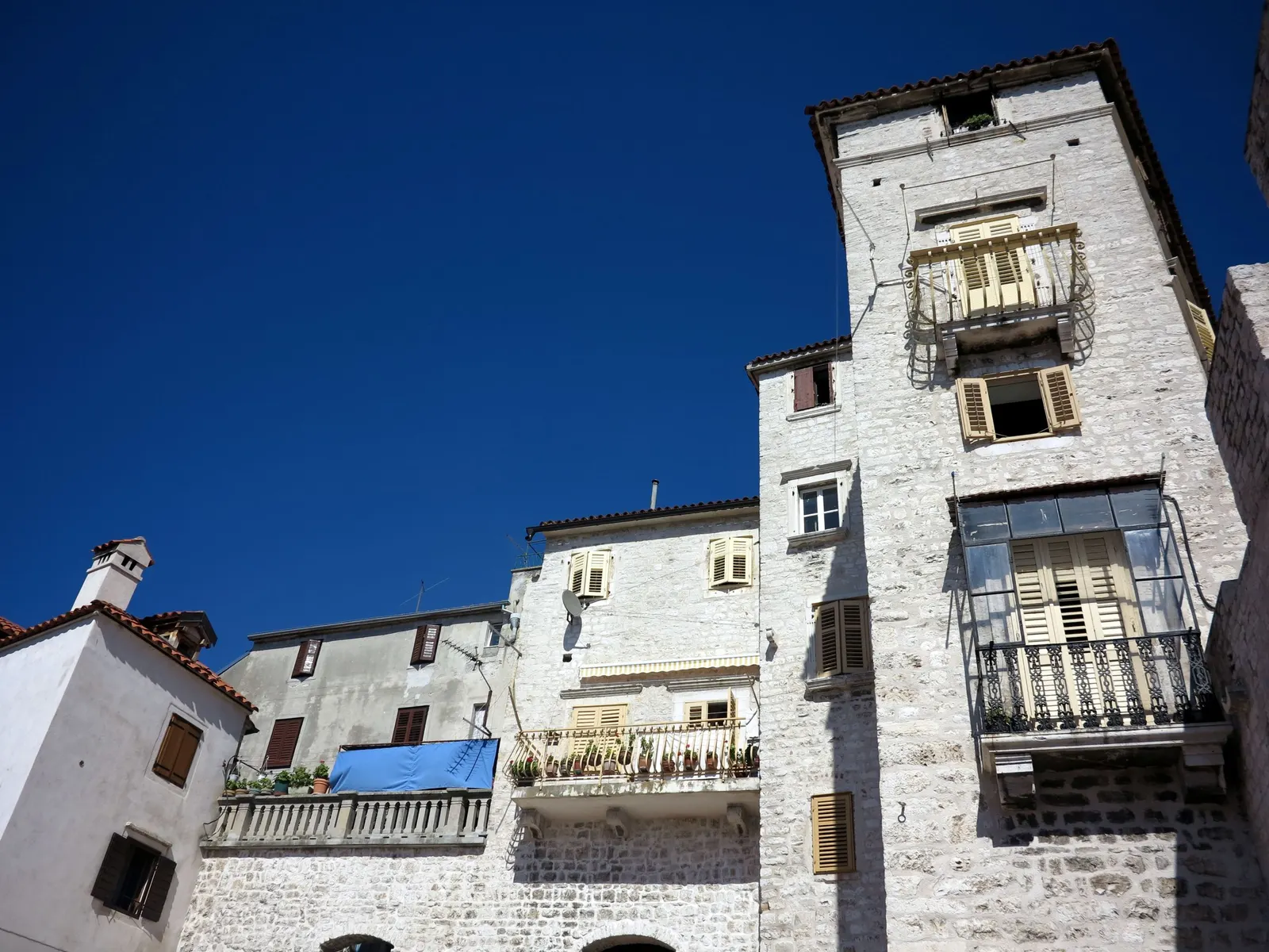 Witte stenen huizen met luiken en balkons in de oude stad van Šibenik onder een strakblauwe lucht