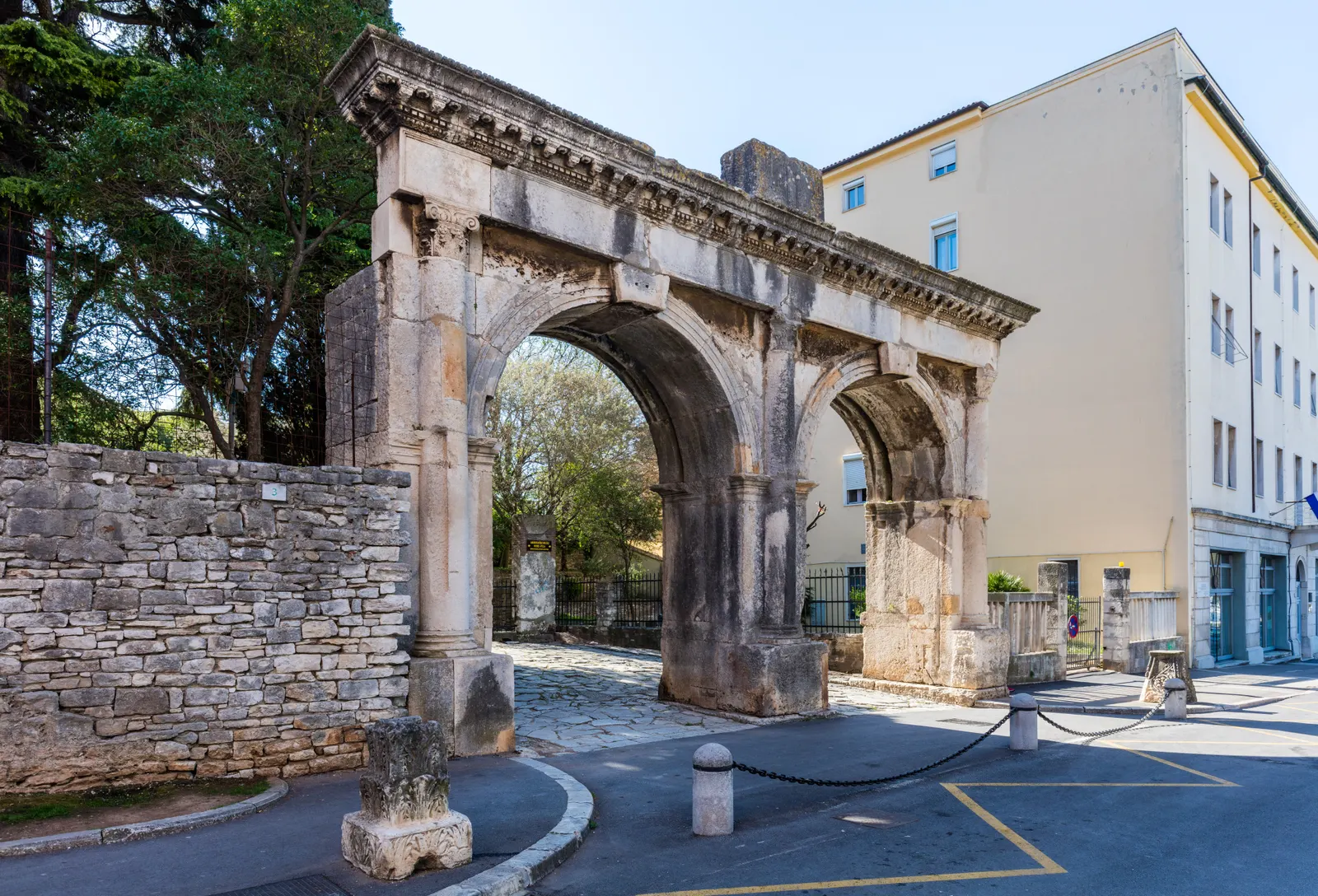 De Romeinse dubbele poort van Pula met twee bogen naast een oude stenen muur en een geel gebouw