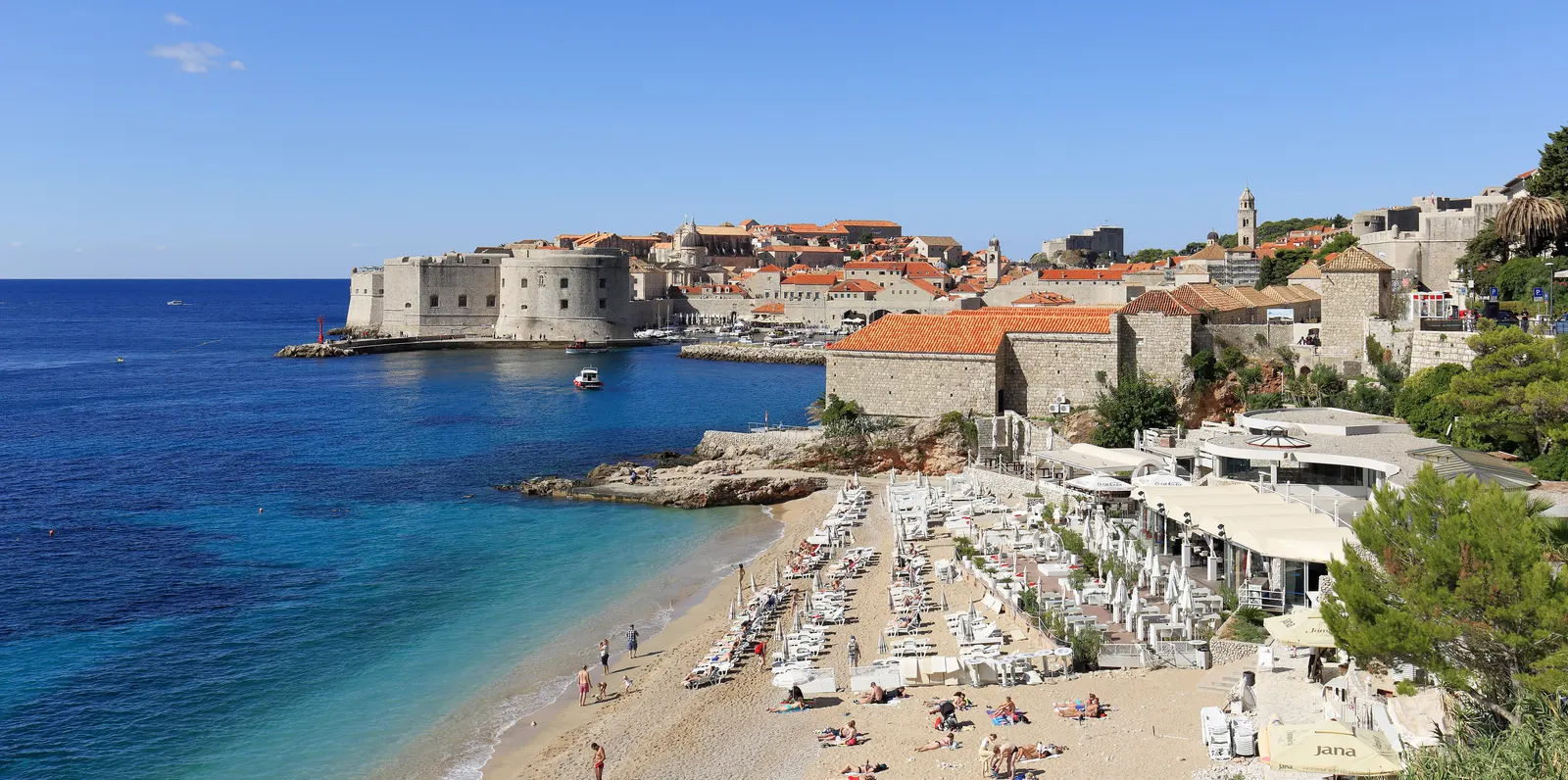 Banje-strand met ligbedden en turquoise zee, met op de achtergrond de stadsmuren van de oude stad van Dubrovnik