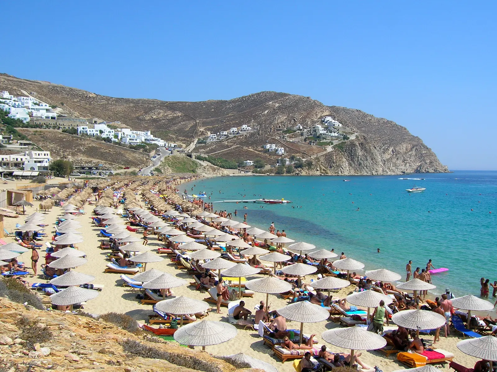 Elia Beach op Mykonos met rijen parasols, turquoise zee en witte gebouwen op de heuvel
