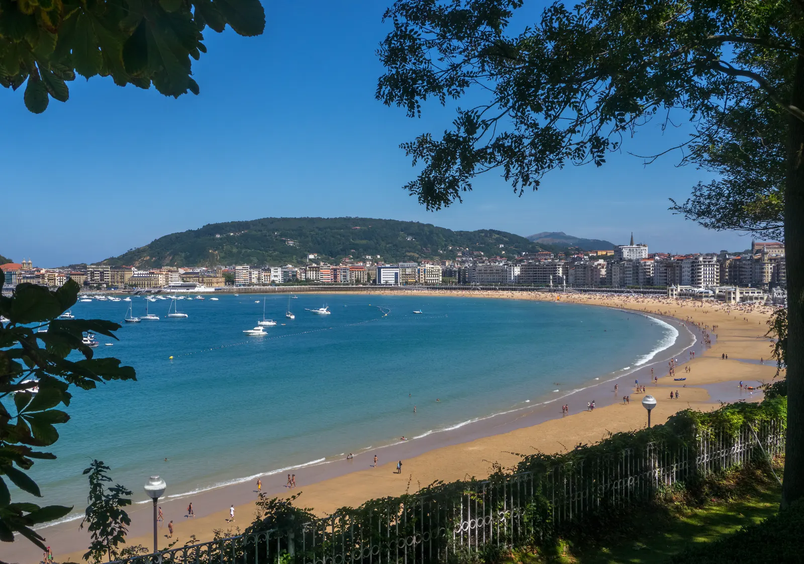 Zonnige baai La Concha met goudkleurig strand, zeilboten op turquoise water en de stad San Sebastian