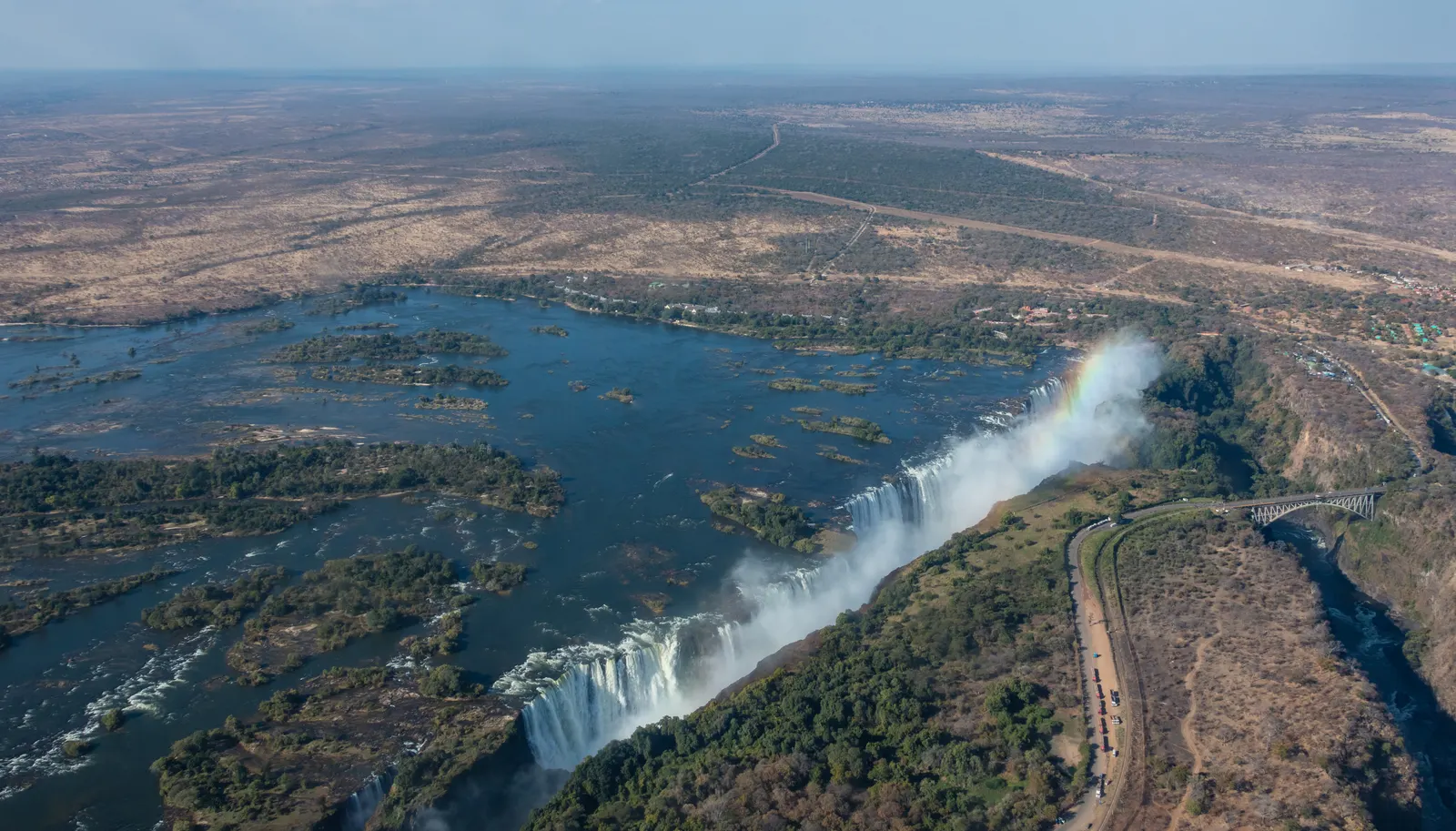 Luchtfoto van de Victoriawatervallen met regenboog in de nevel, de Zambezi-rivier en de Victoria Falls-brug