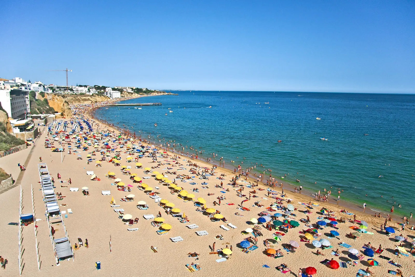 Druk zandstrand met kleurrijke parasols langs helderblauwe zee bij Albufeira, met kliffen op de achtergrond