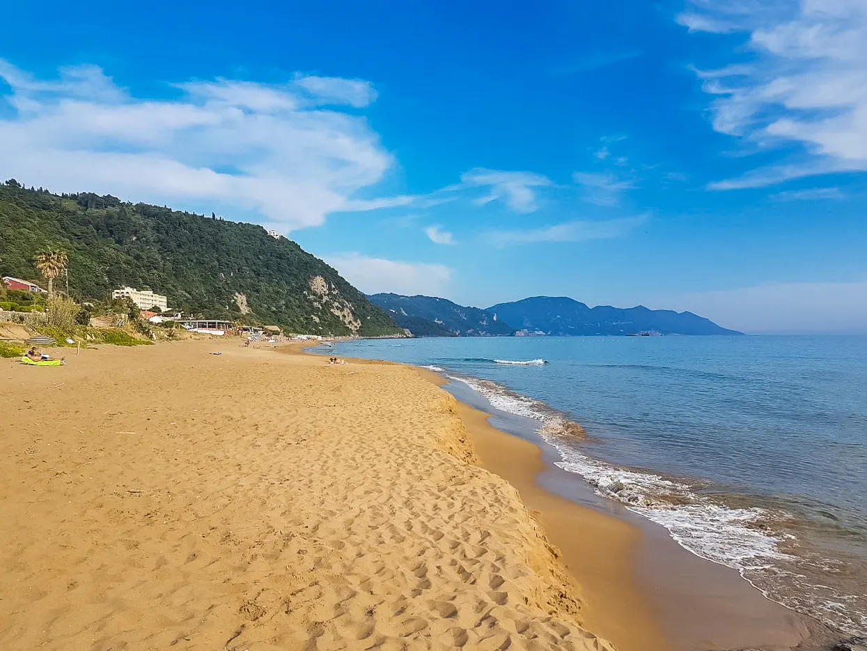 Breed goudgeel zandstrand van Glyfada met golven, groene heuvels en blauwe lucht op Corfu