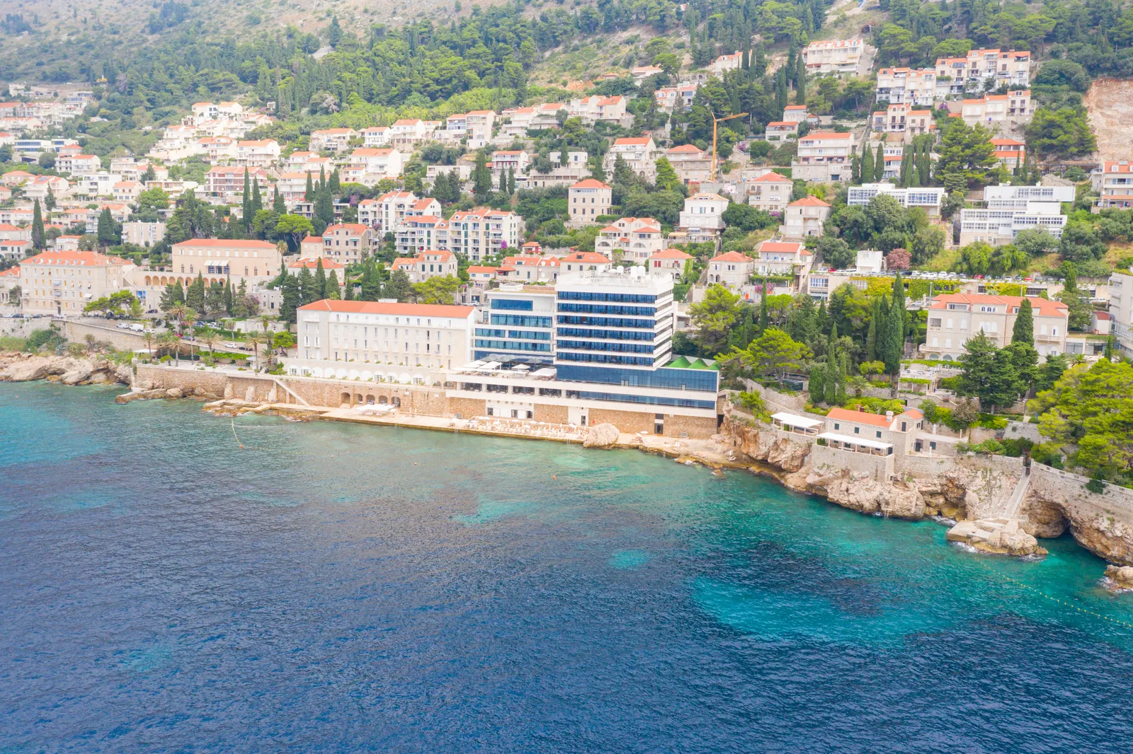 Luchtfoto van Hotel Excelsior aan de rotsachtige kust van Dubrovnik met helderblauwe zee en heuvels vol huizen