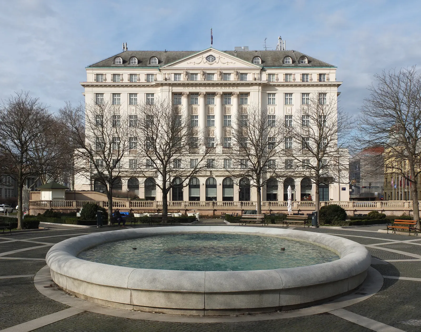 Het witte Esplanade Hotel in Zagreb achter kale bomen, met een ronde fontein op het plein ervoor