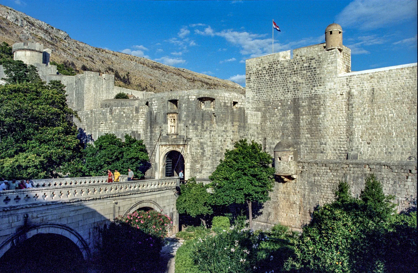 De Pile-poort van Dubrovnik met stenen brug, stadsmuren en groen onder een blauwe lucht