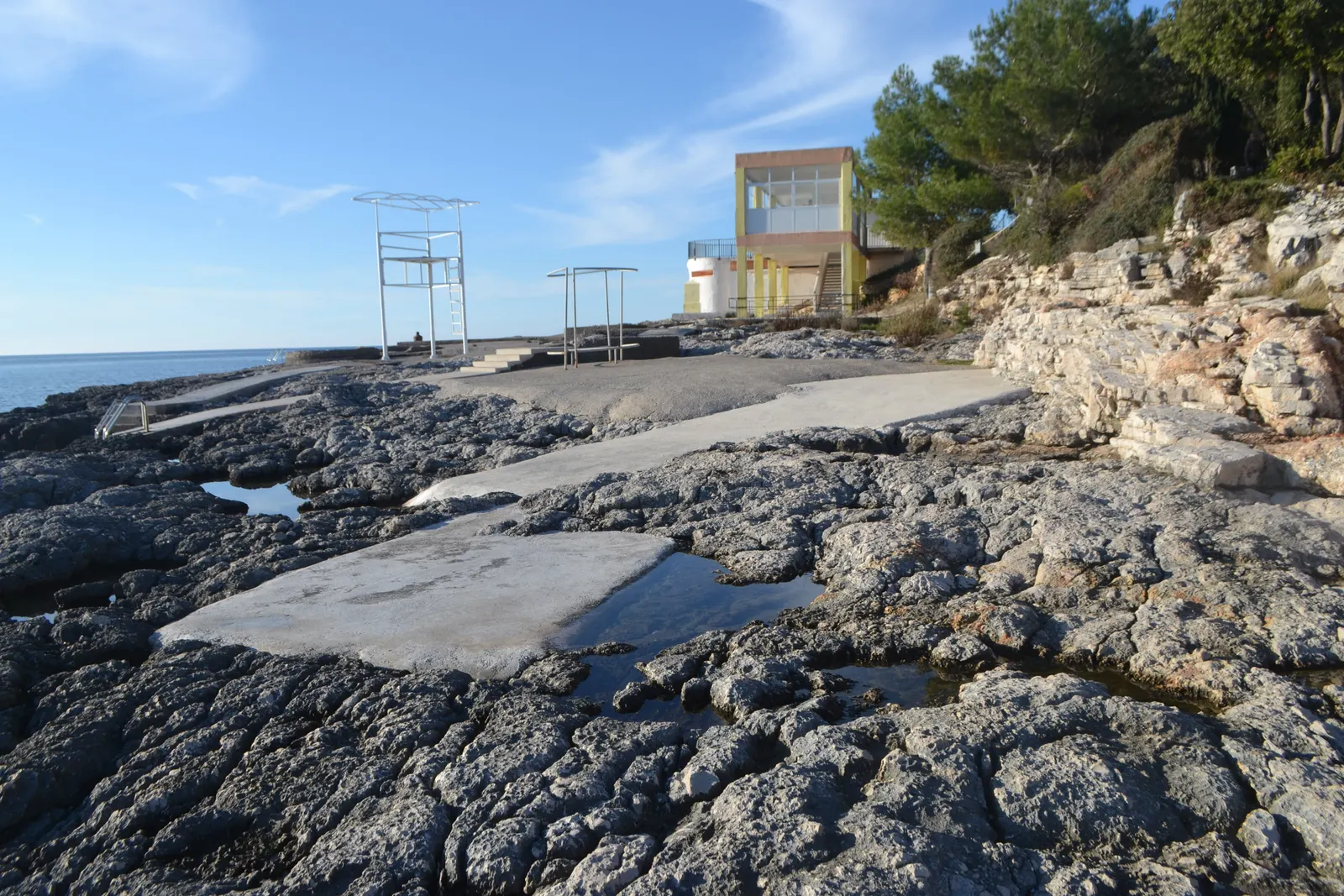 Rotsachtige kust met betonnen ligplekken, zee en een geel gebouw tussen dennenbomen bij Plaza Histria in Pula